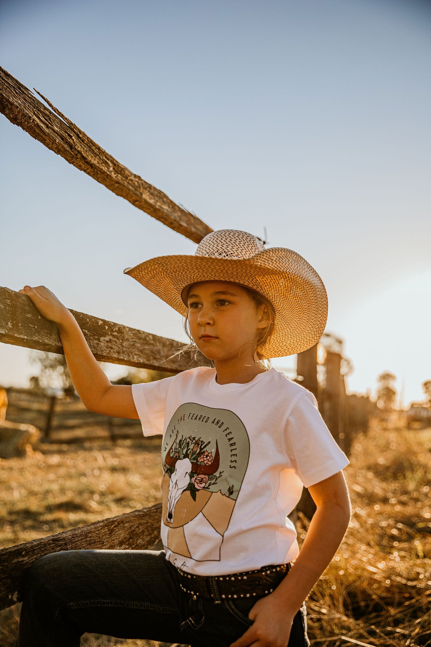 yong youth cowgirl kid wearing  we are the feared & fearless unchained eight country and western tee tshirt for kids youth and ladies shirt has flower cowskull with cactus and sunset. Wearing ariat belt in wrangler or mavericks western wear jeans top end bush boutique standing in paddock in rural australia ringers western cinch jeans little windmill clothing co mavericks western kids straw hat ariat kids western belt