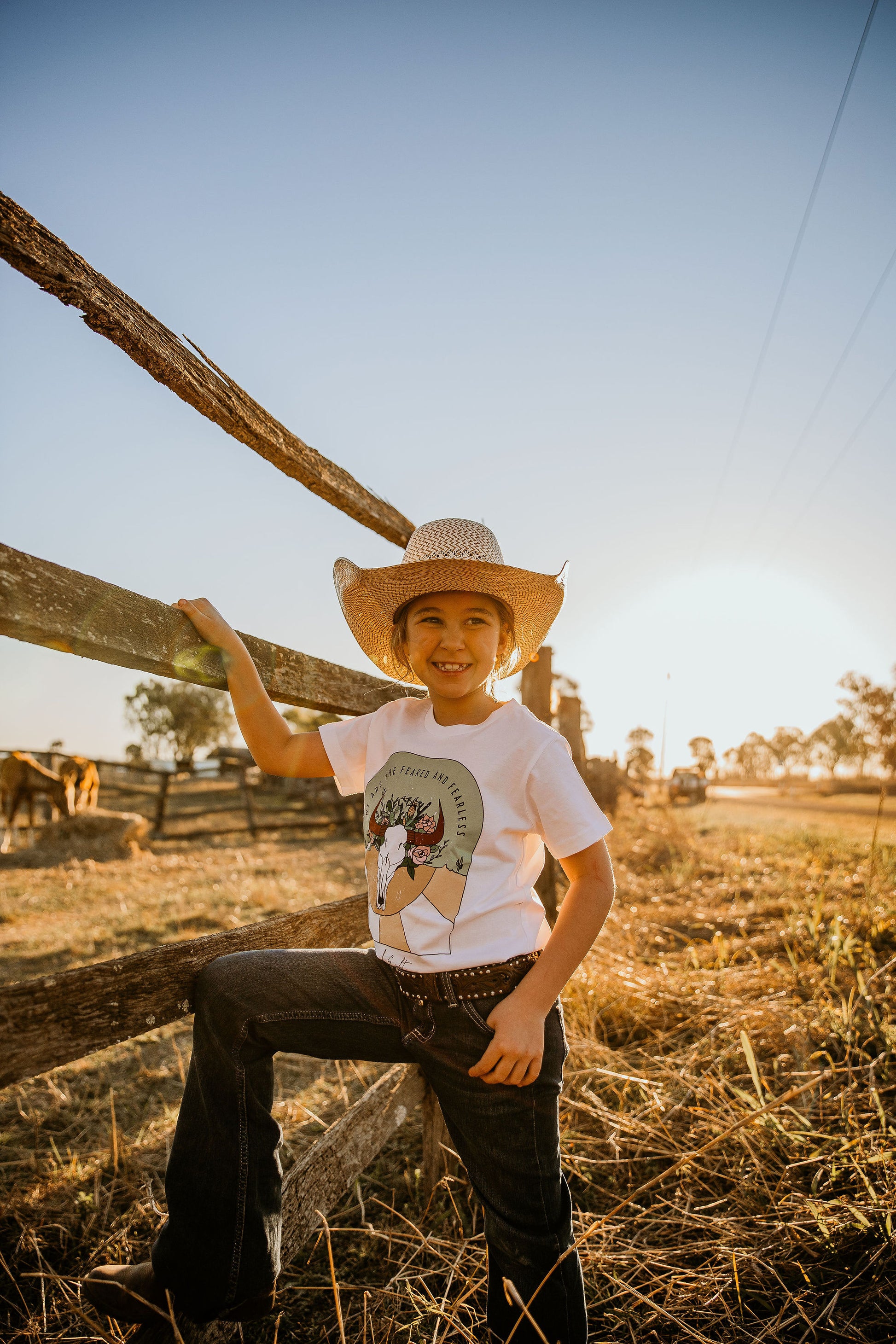 young youth cowgirl kid wearing  we are feared & fearless unchained eight country and western tee tshirt for kids youth and ladies shirt has flower cowskull with cactus and sunset. Wearing ariat belt in wrangler or mavericks western wear jeans top end bush boutique standing in paddock in rural australia ringers western cinch jeans little windmill clothing co australian quarter horse cutting horse campdraft kid in background ariat kids boots circle L kids boots and jeans