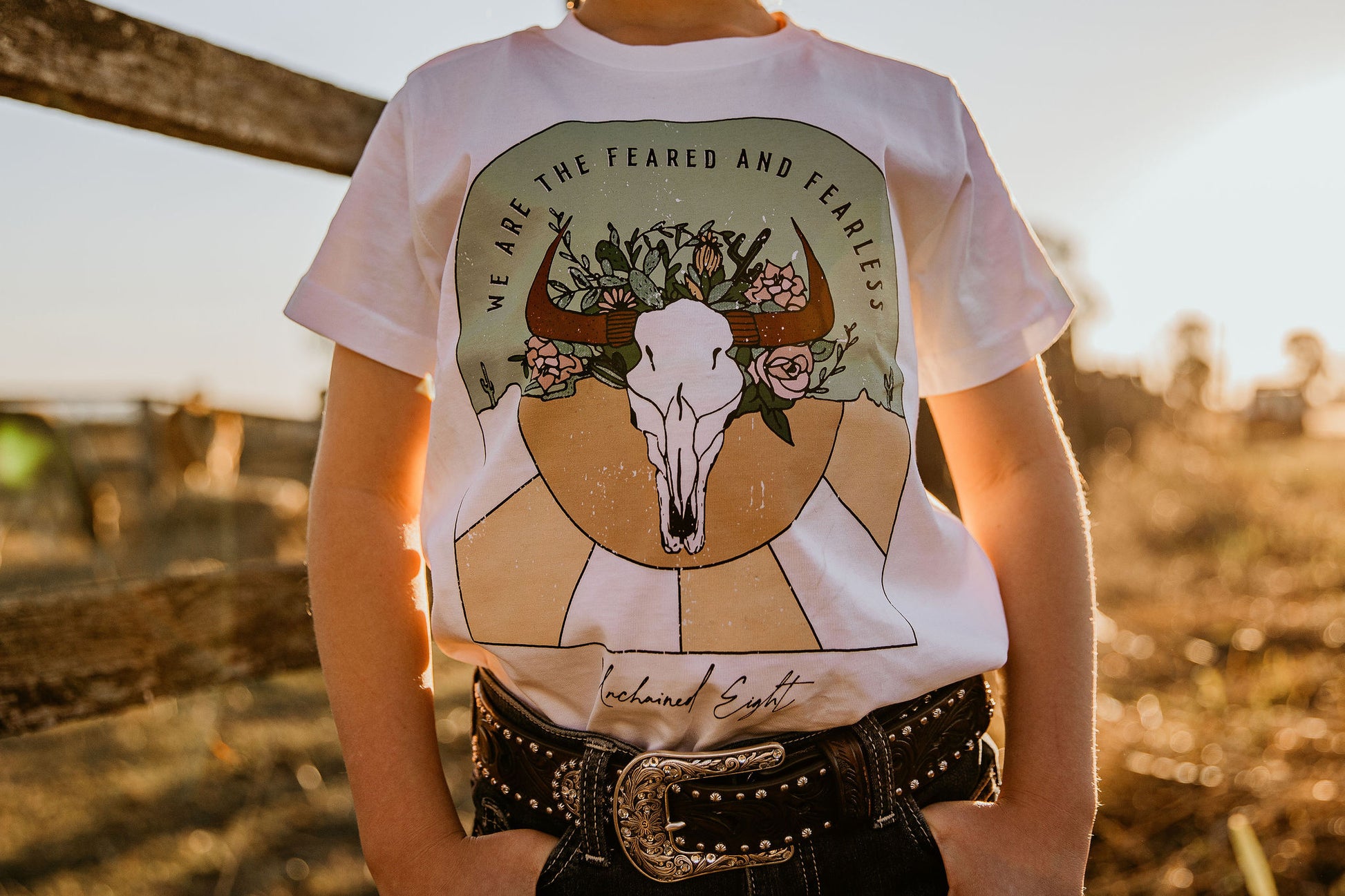 yong youth cowgirl kid wearing  we are the feared & fearless unchained eight country and western tee tshirt for kids youth and ladies shirt has flower cowskull with cactus and sunset. Wearing ariat belt in wrangler or mavericks western wear jeans top end bush boutique standing in paddock in rural australia ringers western cinch jeans little windmill clothing co